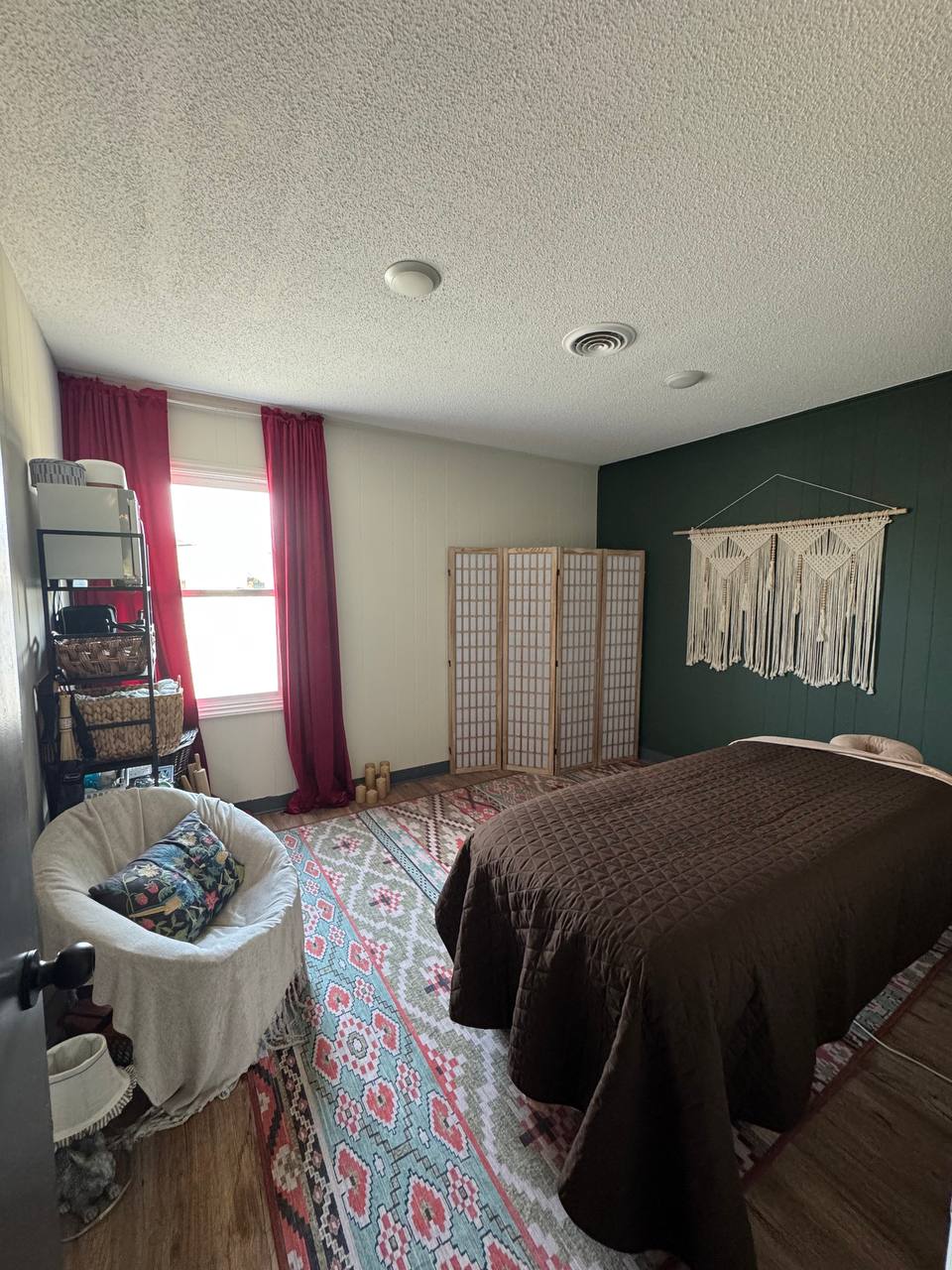 Kira’s studio: a pine green wall with a macrame hanging, crimson curtains, a patterned rug and a massage table